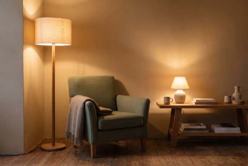 Canto de sala ao entardecer com luz em camadas: candeeiro de pé com abajur de linho e luz de mesa em cerâmica sobre mesa de carvalho