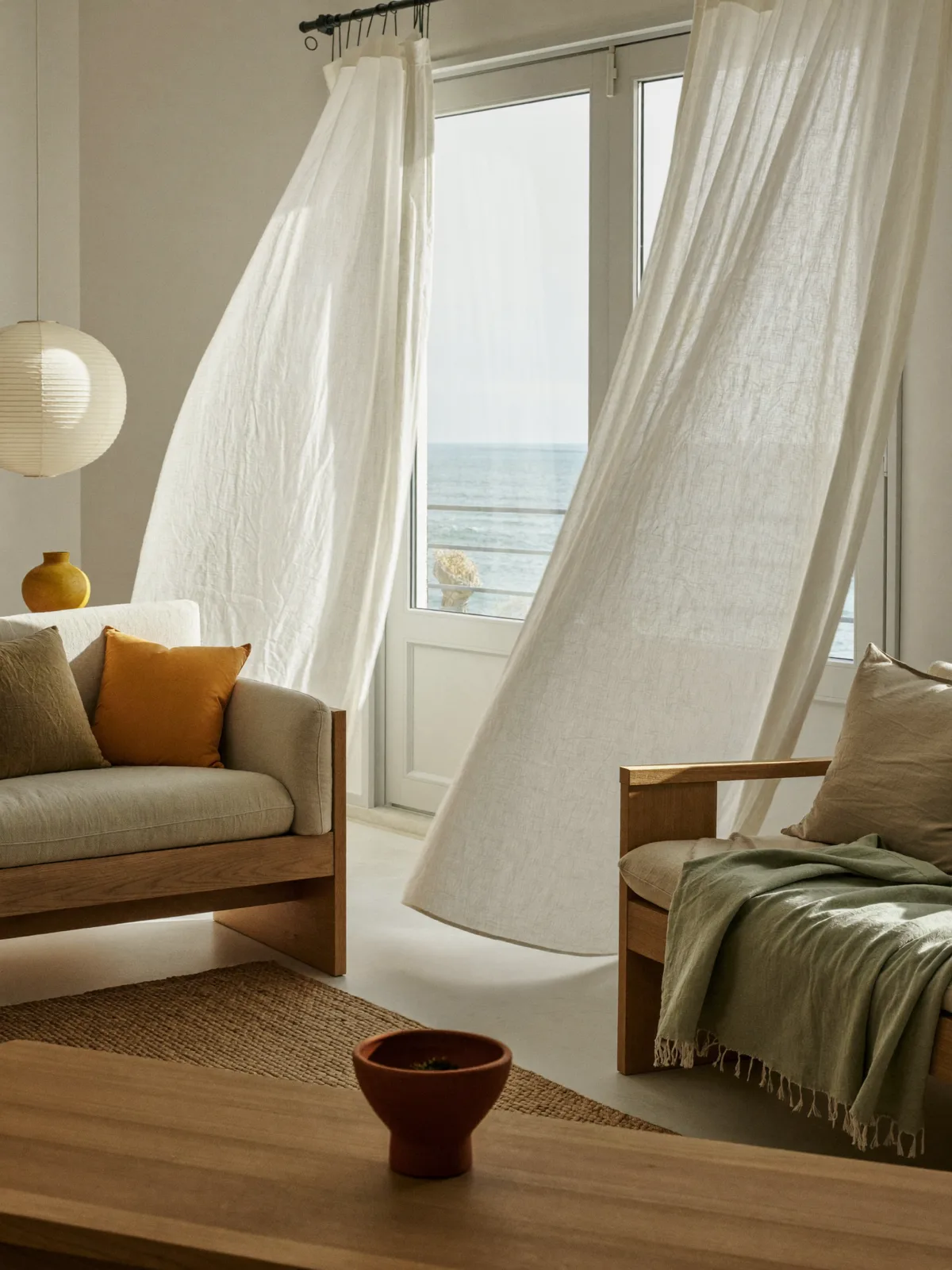 Sala luminosa em estilo mediterrânico: cortinados de linho junto a uma janela alta, madeira clara e tons de areia
